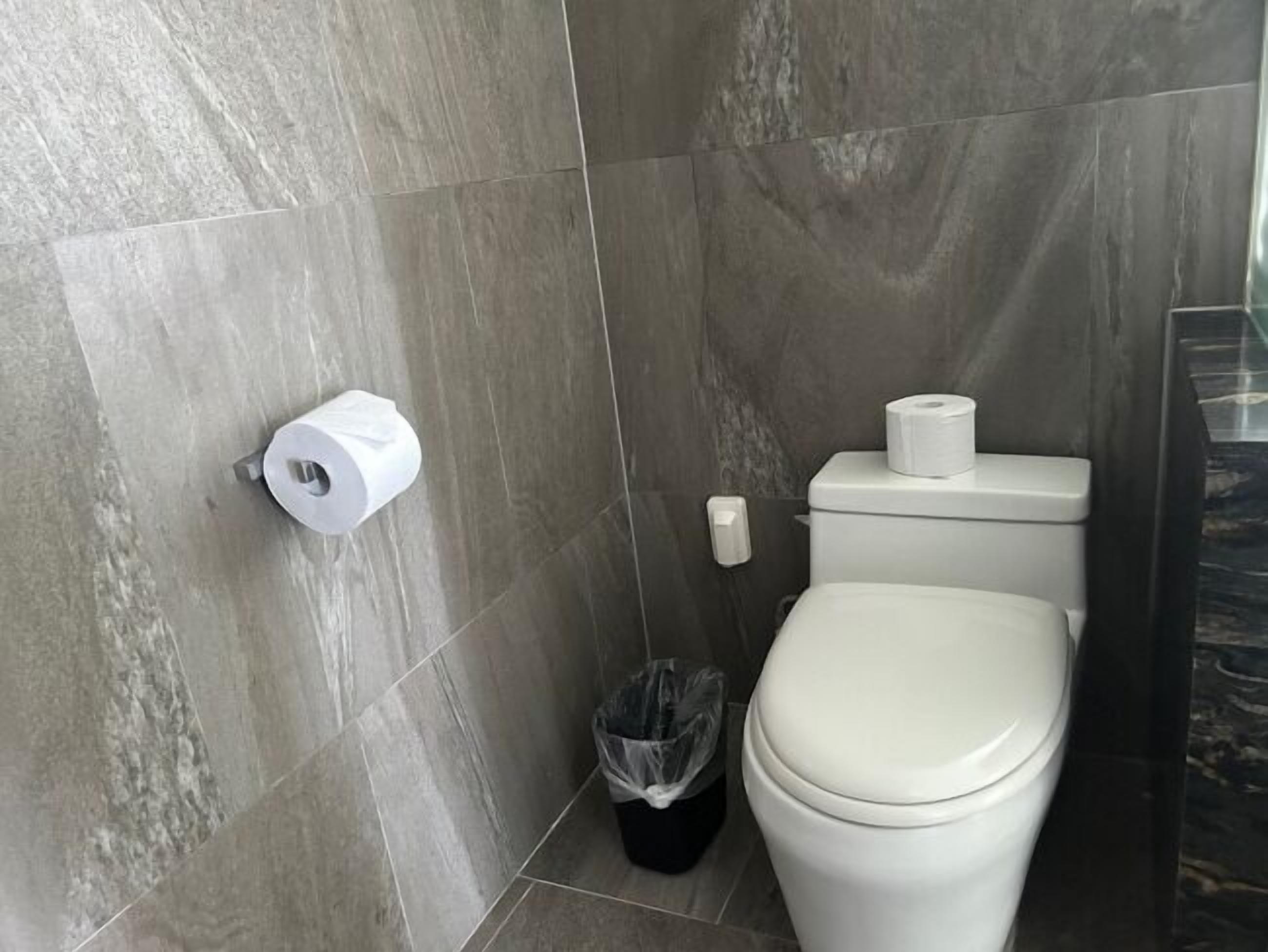 A bathroom with a toilet, a trash bin, and two rolls of toilet paper on the wall.