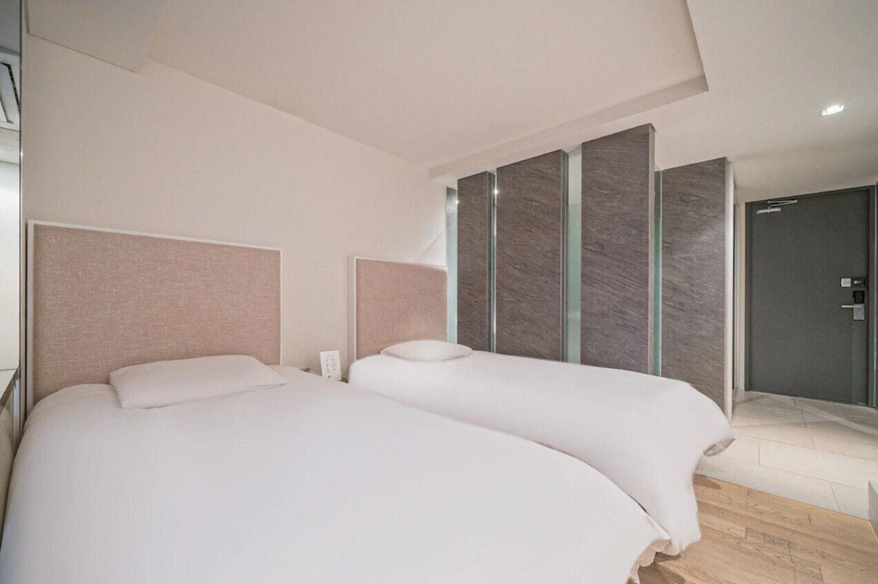 A modern hotel room with two beds, a shower, and a glass partition wall.