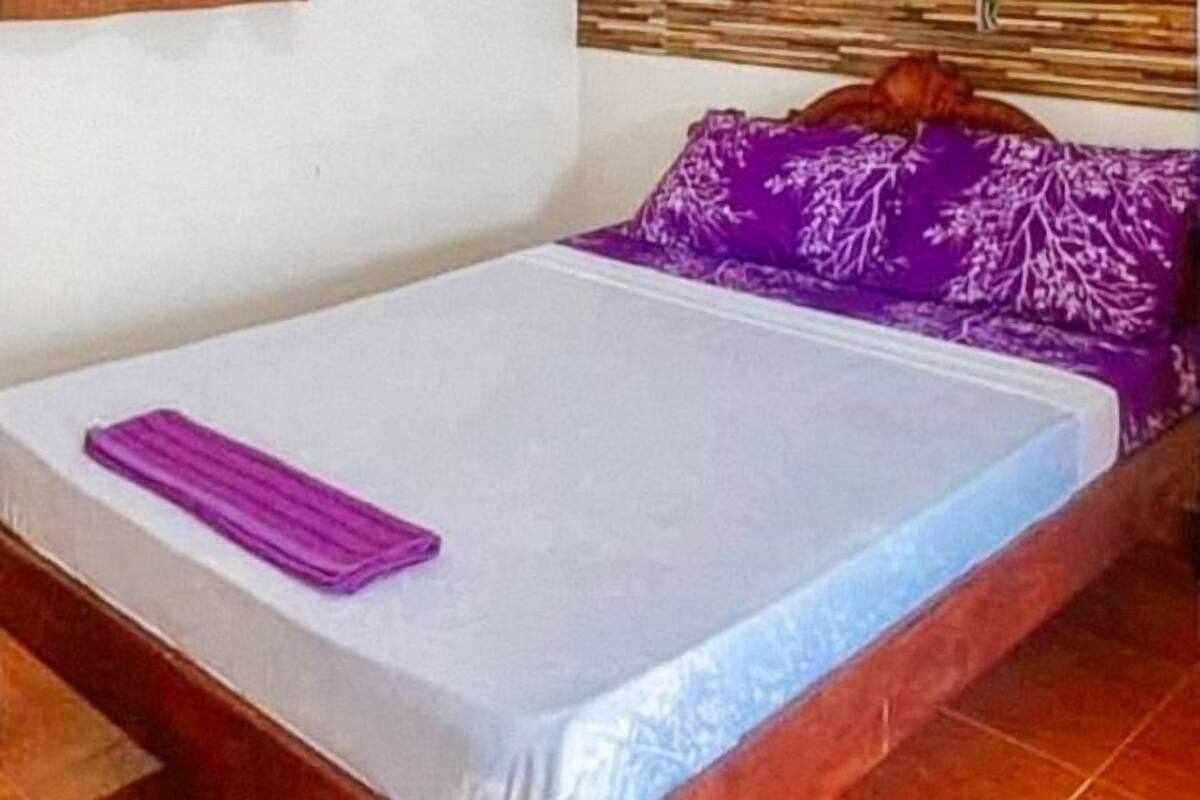 A single bed with a white mattress and a purple blanket, a wooden headboard, and a purple pillow with a leaf pattern.