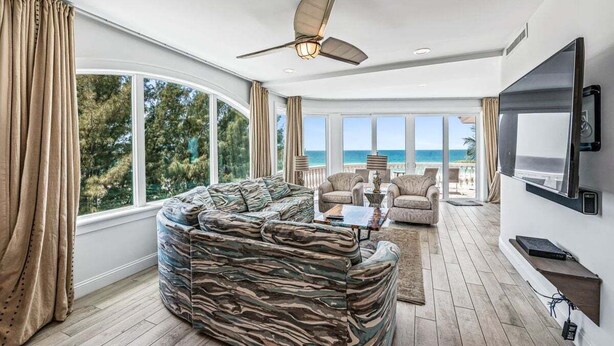 A modern living room with a large curved sofa, a flat-screen TV mounted on the wall, and a ceiling fan. - Anna Maria Grand: Gulf Front! On the Beach! Sleeps 22 by Anna Maria Life Vacation Rentals (Bradenton Beach)