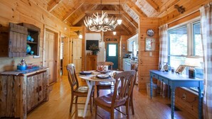 A rustic cabin interior with a wooden dining table, chairs, a chandelier, and a kitchen area visible through an open door. - Annie's Cabin | Pet Friendly | Mountain View | Hot Tub | Fire Pit (Fairview)
