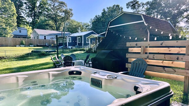 Un bain à remous dans un jardin, avec une terrasse en bois, des chaises et un espace barbecue couvert.