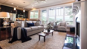 Ein modernes Wohnzimmer mit Sofa, Couchtisch und Sesseln. Es gibt einen Kamin, einen Deckenventilator und große Fenster.