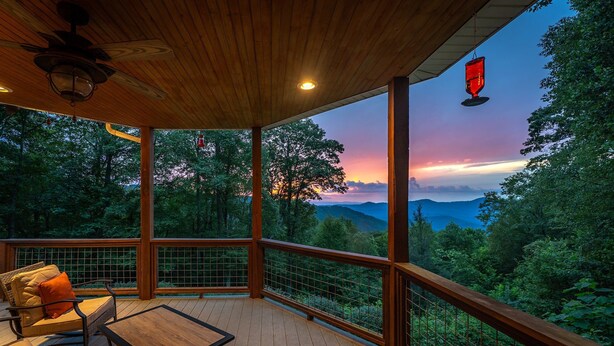 A wooden deck with a table and chairs, overlooking a forested landscape with a sunset view. - Ridgetop Hideaway | Pet-friendly, Hot Tub, Fire Pit & Mountain Views! (Swannanoa)