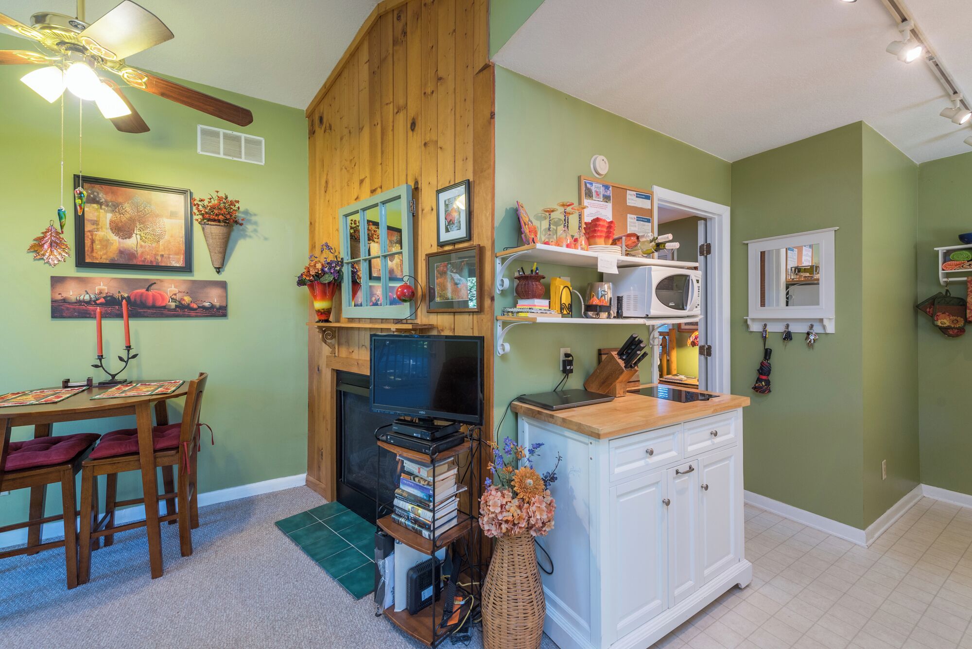 Une pièce chaleureuse avec un ventilateur de plafond en bois, une cheminée, un coin repas avec une table et des chaises, et un coin cuisine équipé d’un four à micro-ondes et de placards.