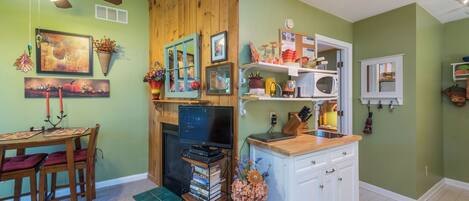 Une pièce chaleureuse avec un ventilateur de plafond en bois, une cheminée, un coin repas avec une table et des chaises, et un coin cuisine équipé d’un four à micro-ondes et de placards.