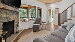 Amplia sala de estar con chimenea de piedra, ventanas grandes, un sofá gris y una escalera de madera.