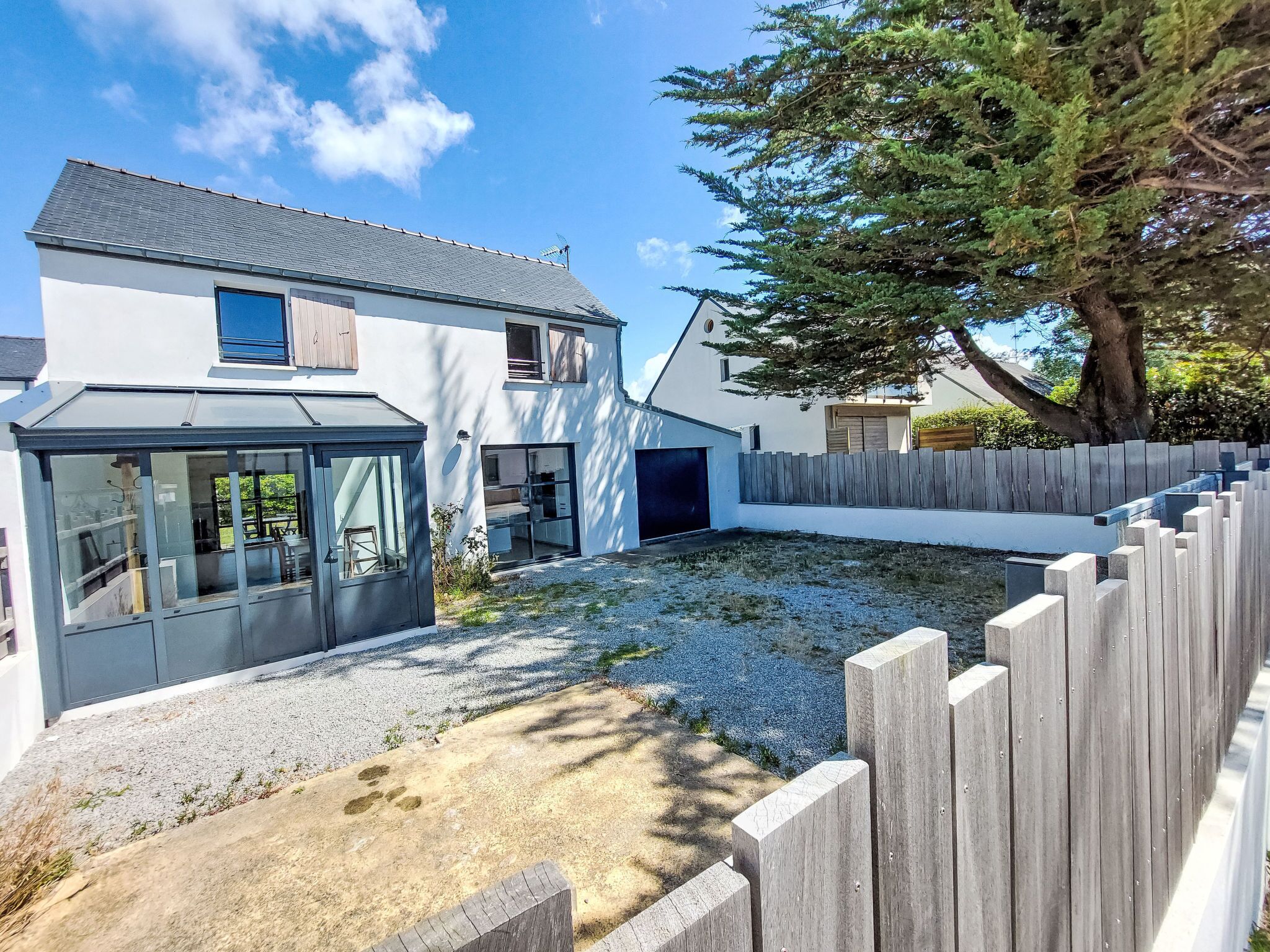 Une maison moderne à deux étages, dotée d’un patio couvert, d’une cour arrière clôturée et d’un grand arbre qui offre de l’ombre.