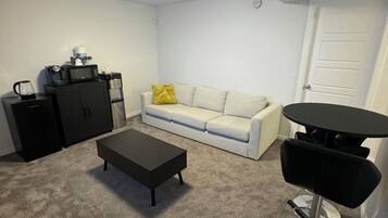 A living room with a grey carpet, a white sofa with a yellow pillow, a black coffee table, a black cabinet, and a black desk with a chair.