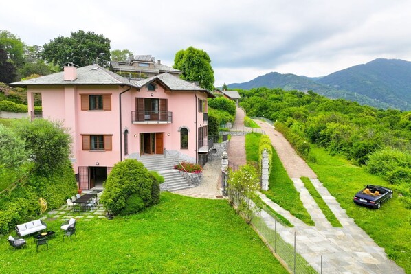 Una casa rosa con una escalera que lleva a un porche, rodeada de abundante vegetación y con montañas al fondo.
