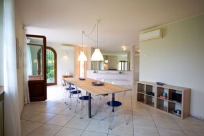Un comedor con mesa y sillas de madera, luces colgantes y una cocina al fondo.