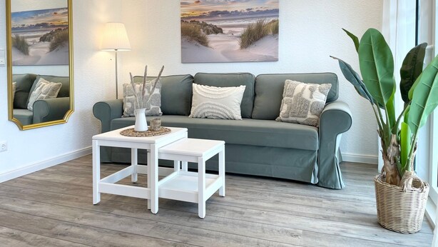 A living room with a grey sofa, white side tables, and two landscape paintings on the wall. - NB 3 - Stylish vacation apartment with private sauna and whirlpool bath - House (Cuxhaven)