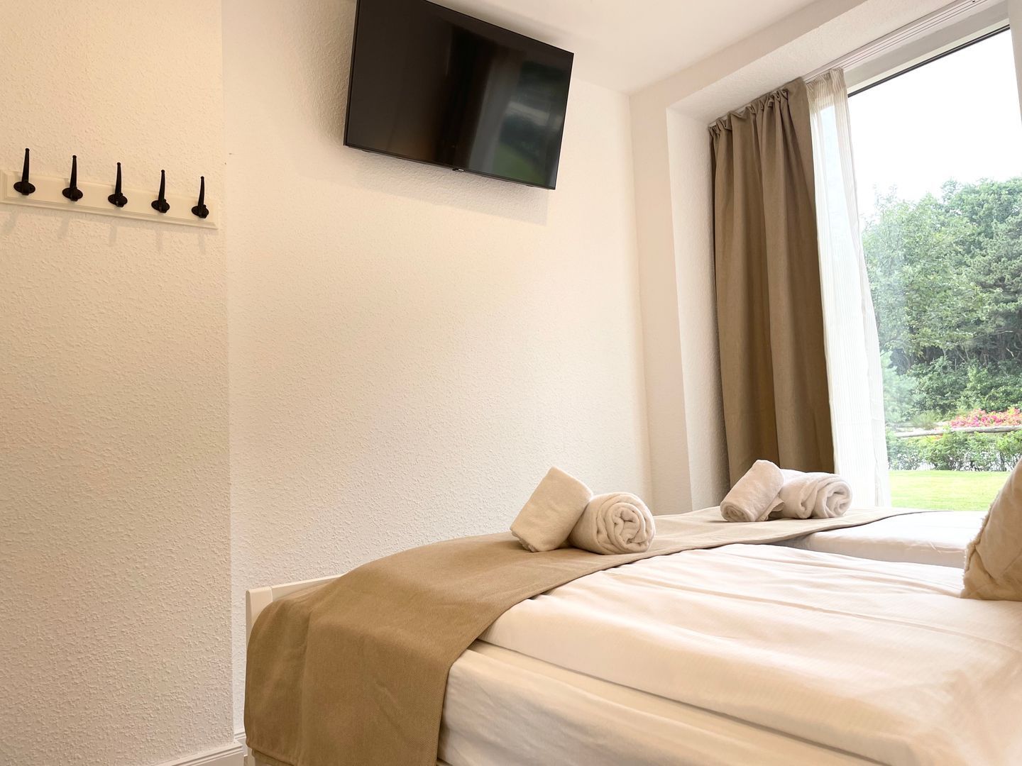A hotel room with a bed, a flat-screen TV mounted on the wall, a window with curtains, and a view of greenery outside.