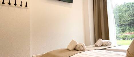 A hotel room with a bed, a flat-screen TV mounted on the wall, a window with curtains, and a view of greenery outside.