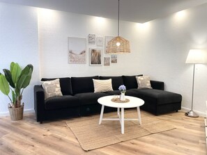A living room with a black sofa, a white coffee table, a hanging light fixture, and a floor lamp. - NB 5 - Strandnahe Ferienwohnung mit Sauna und Whirlpool-badewanne - Haus Nordseebrandung (Cuxhaven)