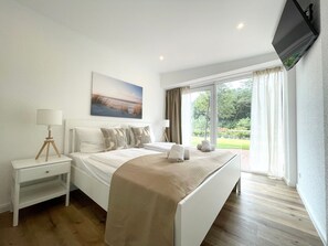 Quarto de hotel com cama, mesas de cabeceira, televisão e uma janela grande com vista para áreas verdes.