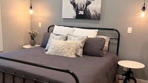 A bedroom with a bed, a bedside table, a wall-mounted artwork, and a fluffy rug. - Spring Branch Cottages "Texas Redbud" (Comal County)