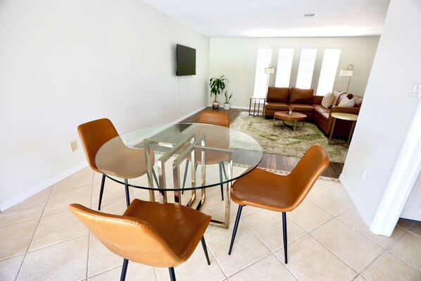 A modern living room with a glass dining table surrounded by four brown chairs, a brown leather sofa, and a small round table. - Ellis' Hideaway! Cozy 2-bedroom pool home with WiFi, AC in enjoyable Cape Coral (Cape Coral)