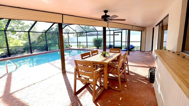 A sunlit patio with a pool, wooden table, and chairs, overlooking a scenic view.
