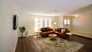 A living room with a brown sectional sofa, a coffee table, and a TV mounted on the wall. There are two potted plants and a rug on the floor.