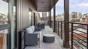 Ein Balkon mit einer Couch, Hocker und Stühlen mit Blick auf eine Stadtlandschaft.