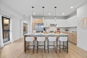 Una cocina moderna con una isla central, taburetes de bar, iluminación colgante y vistas al área de estar.