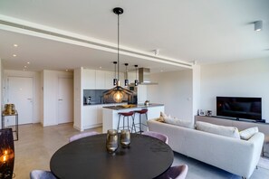 A modern living room with a round dining table, a sofa, a TV, and a kitchen area with bar stools.