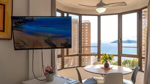 Une pièce dotée d’un téléviseur diffusant des images d’une plage, d’une table à manger avec des chaises, et d’une grande fenêtre offrant une vue sur les bâtiments et la mer.