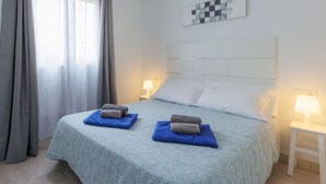 Ein Hotelzimmer mit einem Bett, zwei Kissen, einer Decke, einem Nachttisch, einer Lampe und einem Fenster mit Vorhängen.