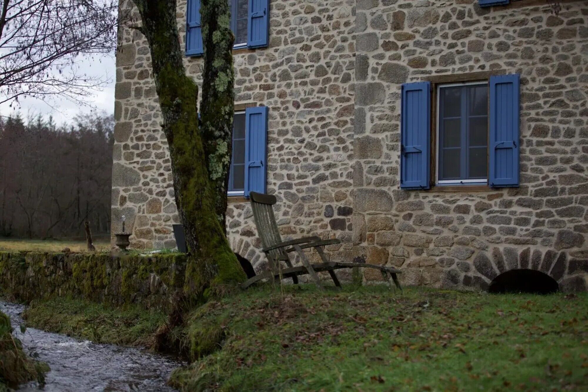 Een stenen gebouw met blauwe luiken, een rustieke bank en een klein beekje.