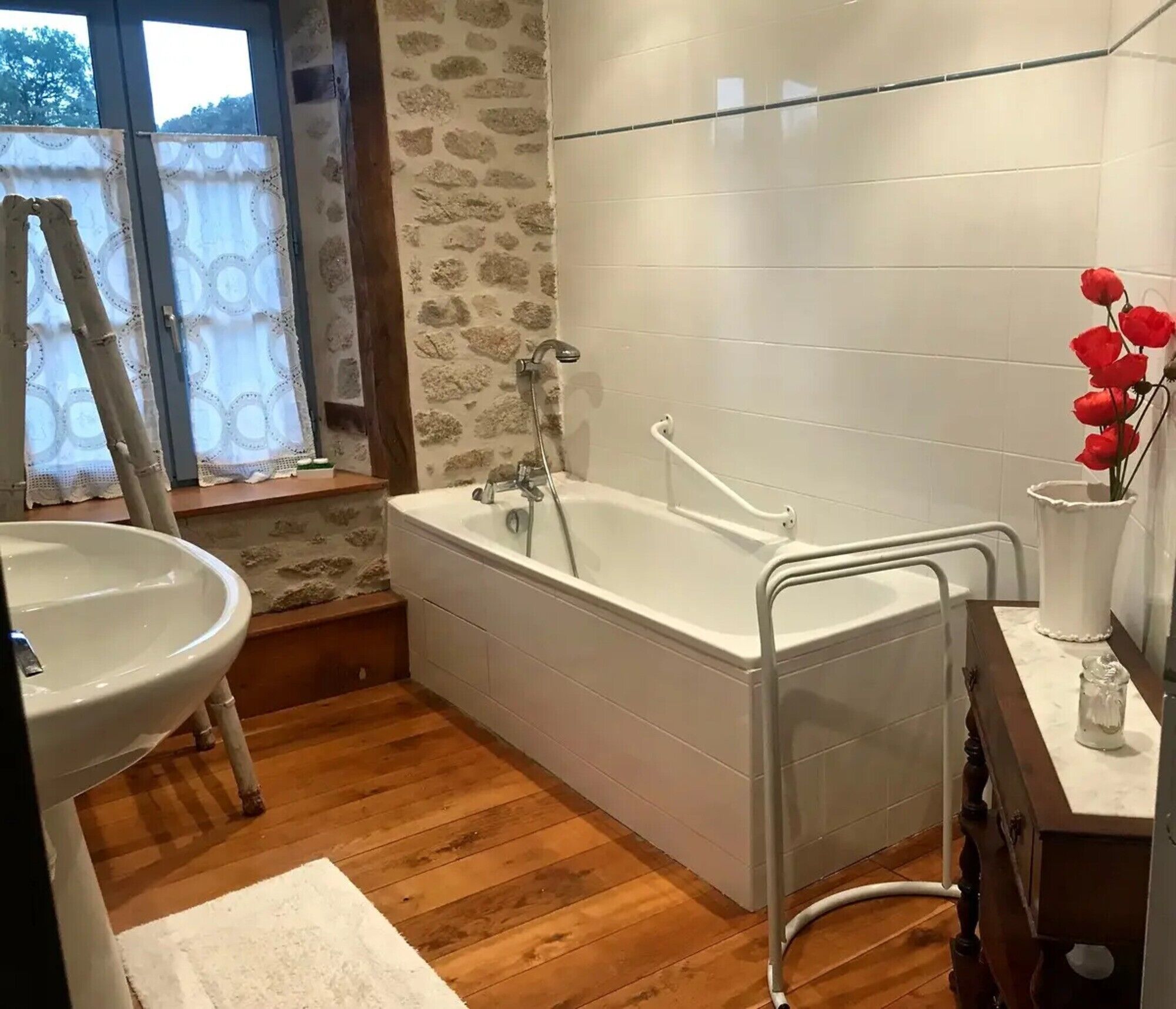 A bathroom with a bathtub, a sink, a window with curtains, and a vase with flowers.
