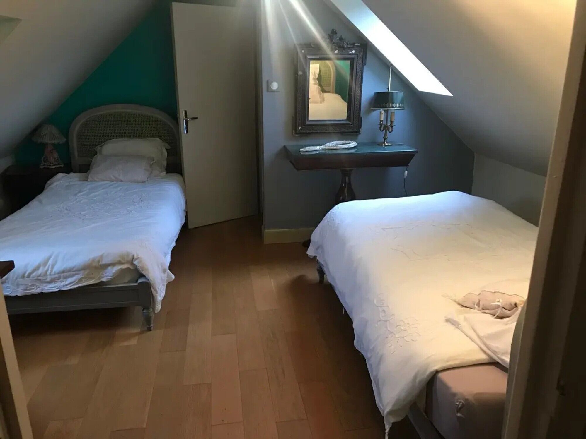 Una habitación pequeña y acogedora con dos camas, suelo de madera, un espejo y una mesita con una lámpara.
