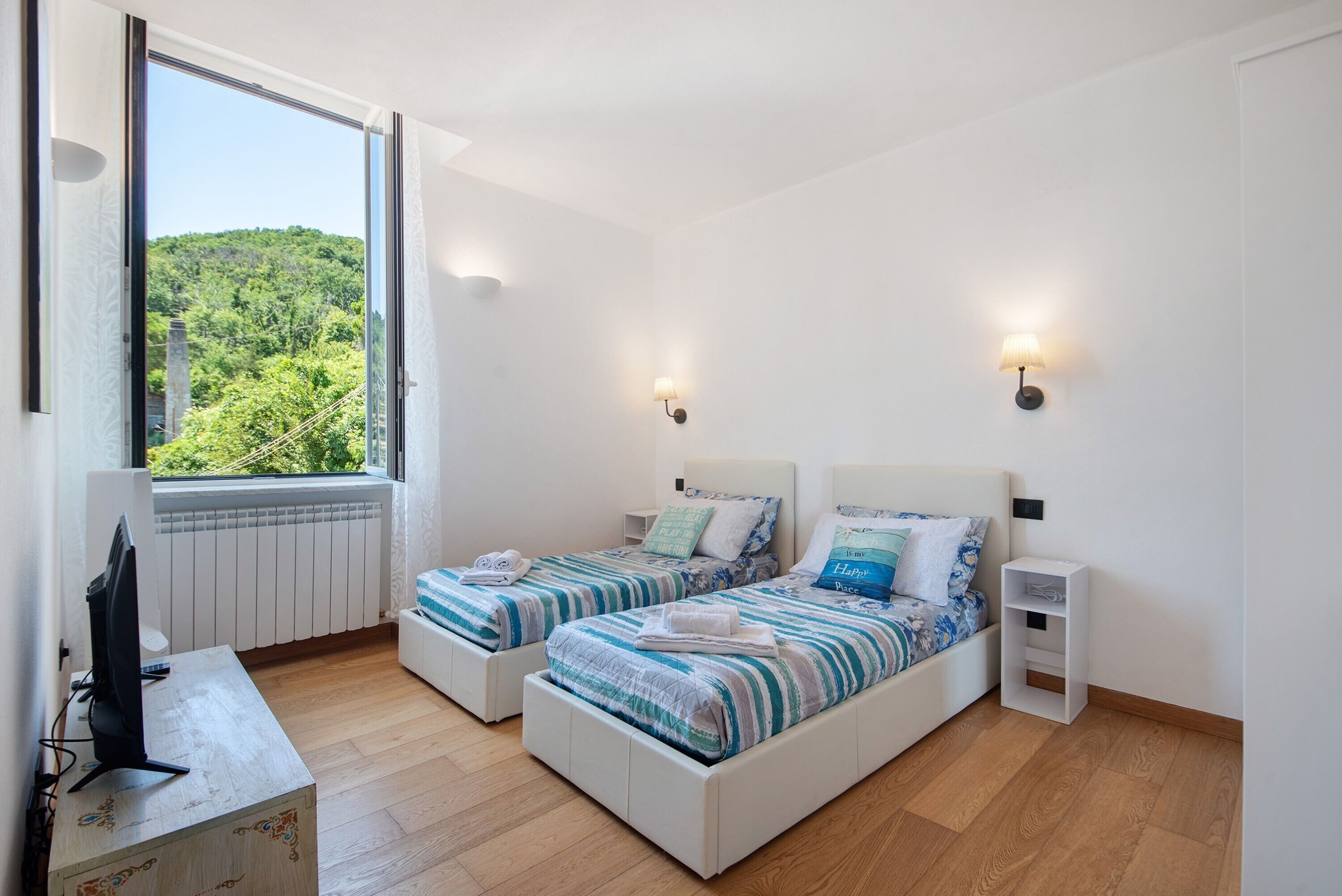 Ein Zimmer mit zwei Betten, einem Schreibtisch, einem Fenster mit Blick auf Grünflächen und Wandleuchten.