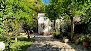 A cobblestone pathway leading to a white building with a covered patio area, surrounded by lush greenery and potted plants.