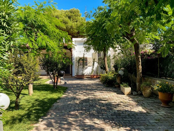 A cobblestone pathway leading to a white building with a covered patio area, surrounded by lush greenery and potted plants.