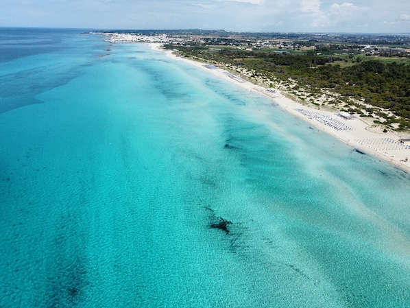 An aerial view of a coastal area with a beach, clear turquoise waters, and a distant shoreline with buildings.