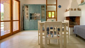 A room with a dining table, chairs, a fireplace, a window with a view, and a kitchen area with cabinets.