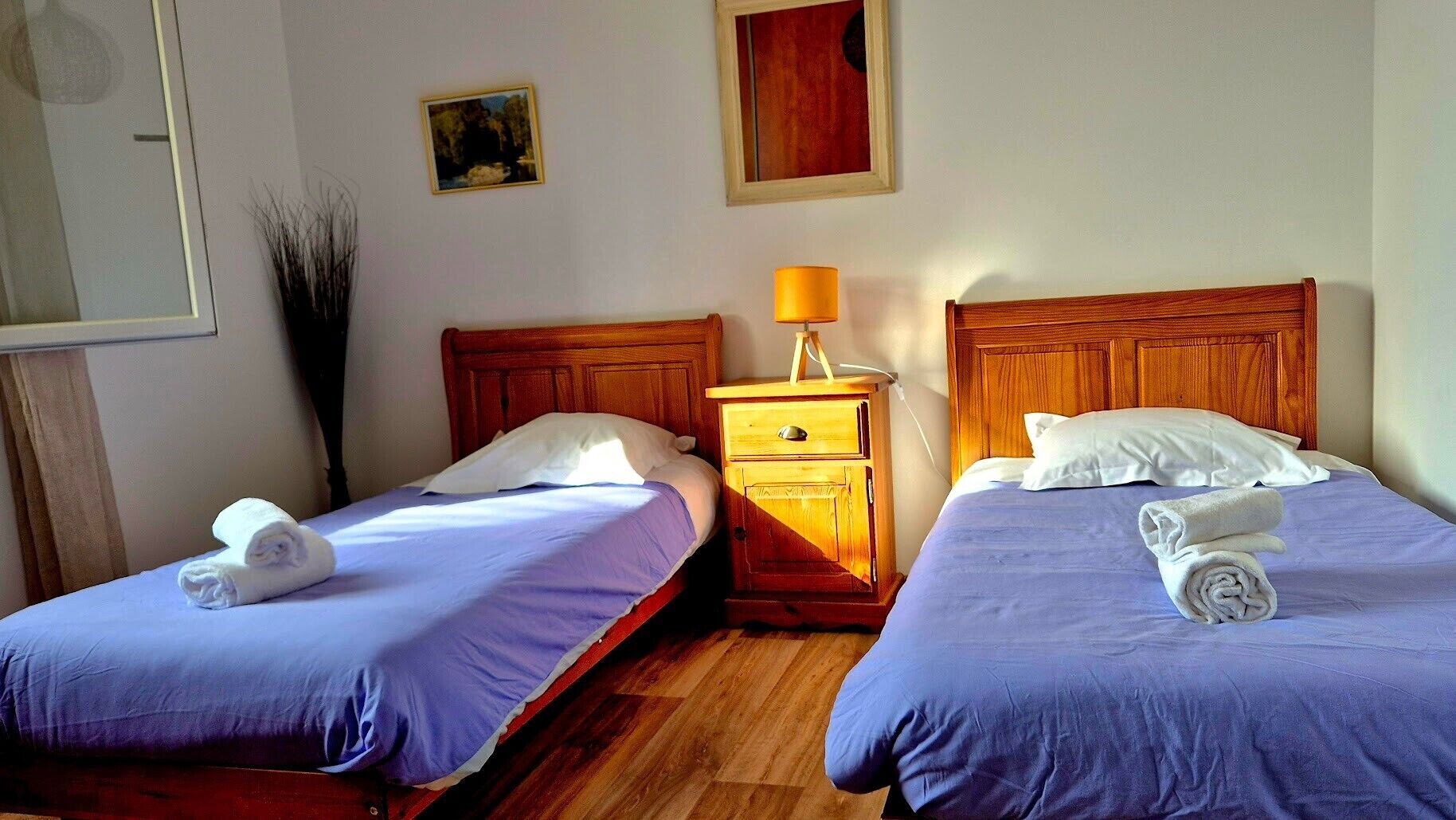 A hotel room with two single beds, wooden headboards, a nightstand with a lamp, and a painting on the wall.