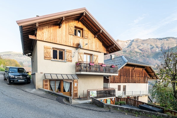 A two-story wooden house with a balcony, a car parked in front, and a mountainous backdrop.