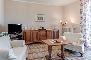 A living room with a television, wooden cabinet, two white armchairs, a coffee table, and a radiator.