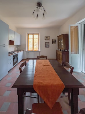A dining area with a long table, chairs, a kitchen with cabinets, a radiator, and a window with curtains.