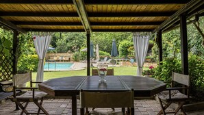 An outdoor patio with a wooden table and chairs, a pool in the background, and lush greenery.