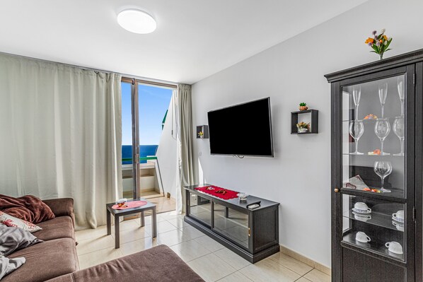 Una sala de estar con un sofá, un televisor, un aparador de vidrio con tazas y vistas al mar.