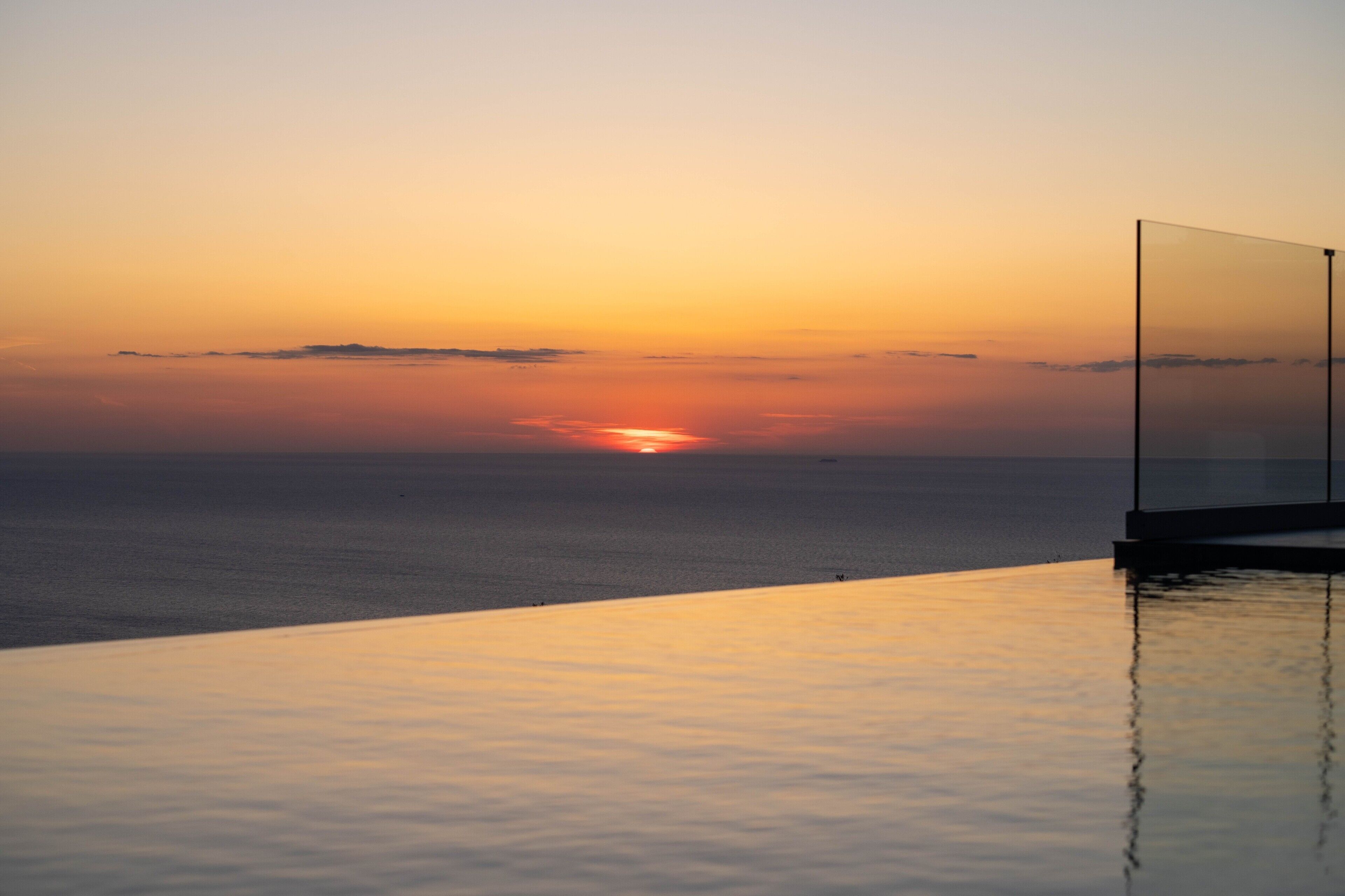 Een zonsondergang boven de oceaan met een heldere lucht, een zwembad en een glazen balustrade.