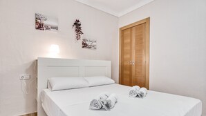 Quarto com uma cama branca, uma porta de madeira e duas imagens penduradas na parede.