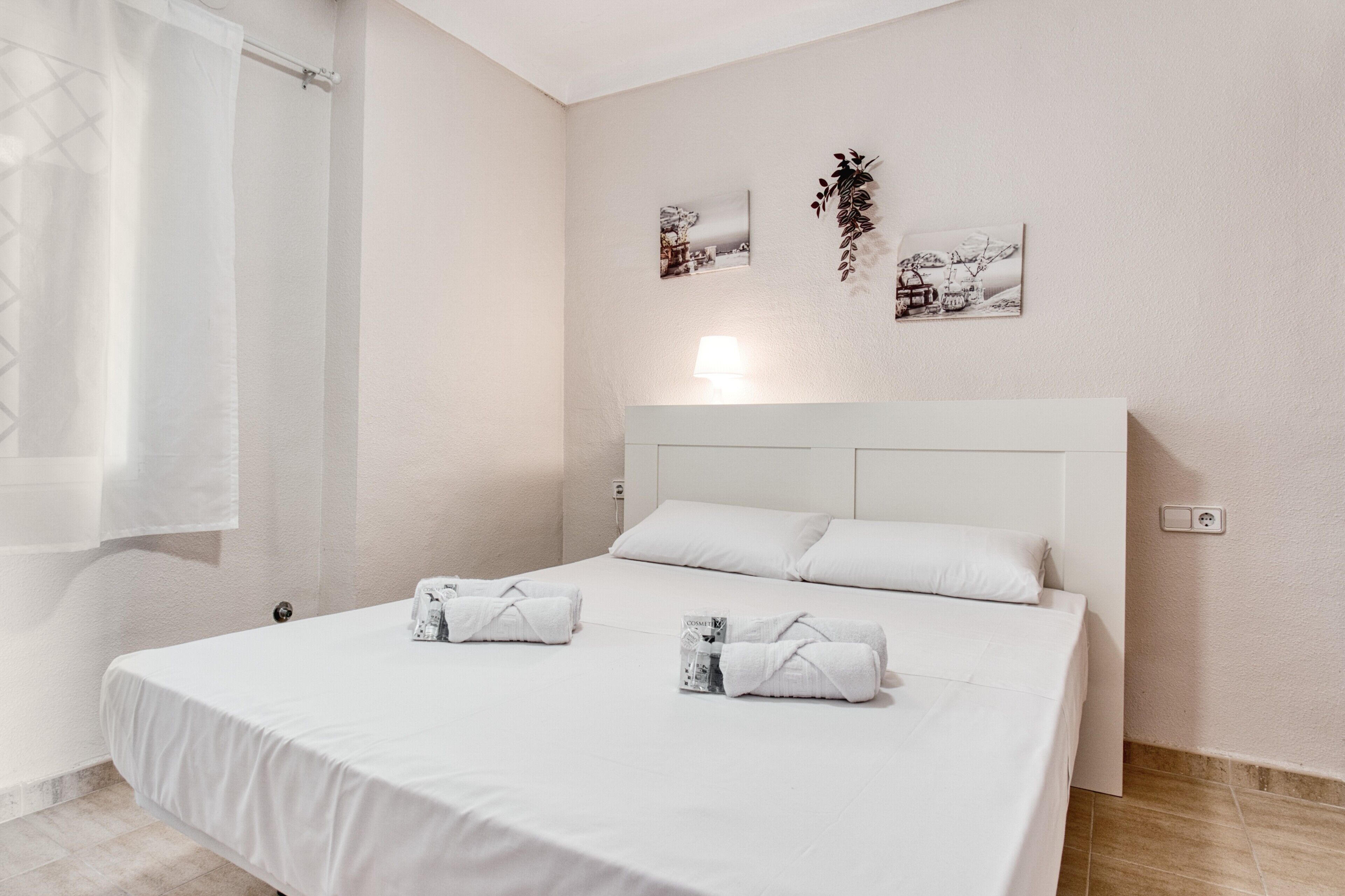 A bedroom with a large bed, white bedding, two rolled towels, a nightstand, a wall-mounted lamp, and two framed pictures.