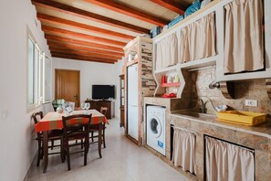 A compact kitchen with a washing machine, a dining area with a table and chairs, and a television in the background.