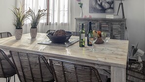 Un coin repas avec une table en marbre, des chaises en osier, un saladier, des bouteilles de vin et un tableau accroché au mur.