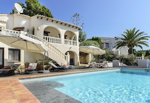 Zona de piscina con sillones de descanso, piscina y un edificio con arcos y un balcón.