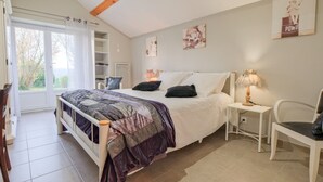 A bedroom with a bed, a bedside table, a chair, a window with curtains, and a view of the outdoors. - Gite Le Moutat-La Cave with Private Garden and Wi-Fi (Allemans)